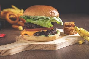 A cheeseburger with lettuce and tomatoes on top of a wooden cutting board with fries and onion rings on the counter in the background. | St. Louis, MO